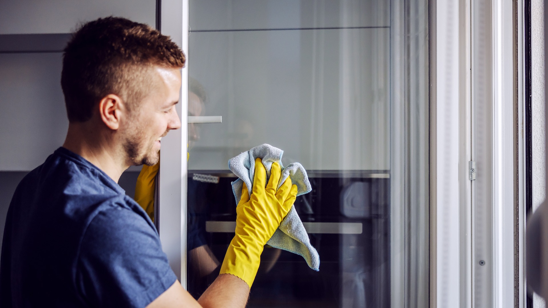 Young bearded smiling positive man with rubber gloves cleaning window with cloth after rain there is always stains on glass jpg